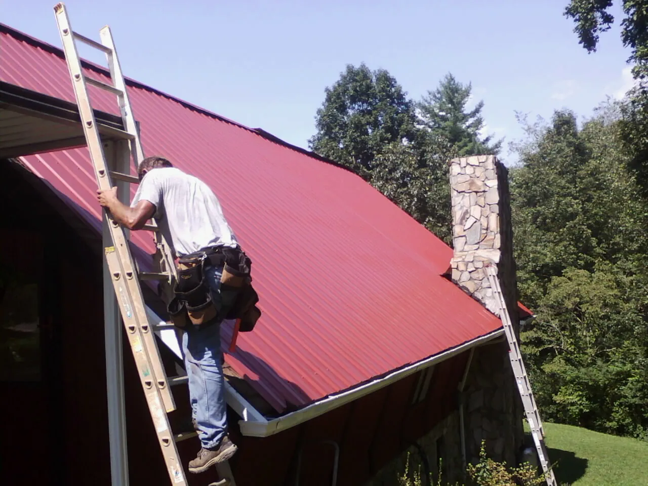 Roofer on a ladder beside a red metal roof and stone chimney — Soffit & Fascia Repair by Roofing Brothers in Perkinsville